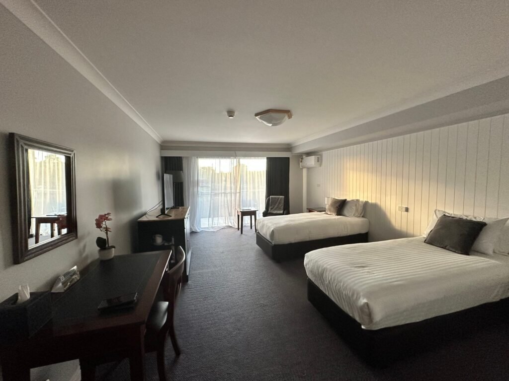 A spacious hotel room with two beds, a desk, and soft light coming through a window